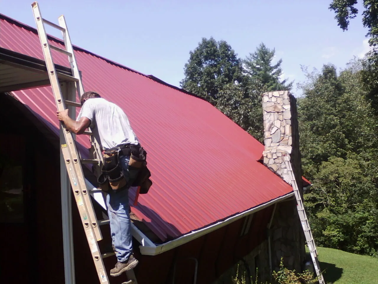 Roofer on a ladder beside a red metal roof and stone chimney — Soffit & Fascia Repair by Roofing Brothers in Collegeville